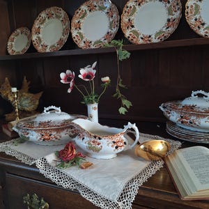 K&ouml;nnte beinhalten: Eine Pr&auml;sentation von antiken Porzellan mit floralen Mustern in Orange, Braun und Gr&uuml;n. Das Set umfasst Teller, Servierschalen und eine Sauciere, die auf einem dunklen Holzschrank angeordnet sind. Ein wei&szlig;es Spitzendeckchen und eine goldfarbene Schale sind ebenfalls vorhanden.