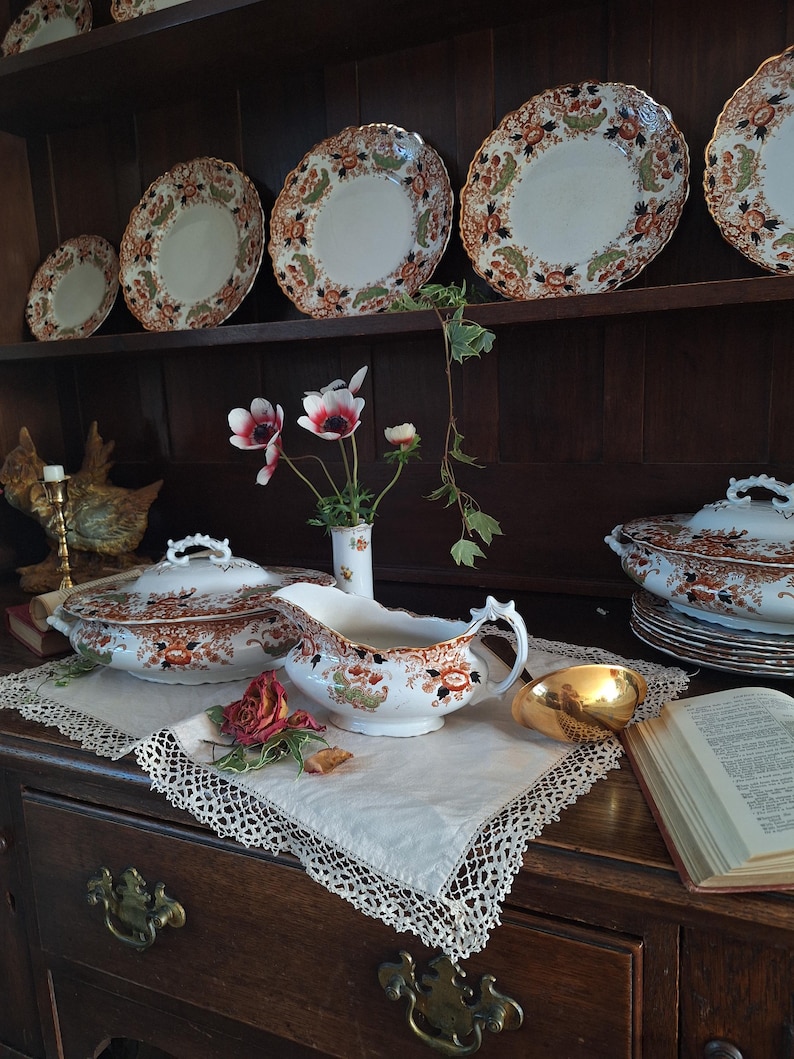 K&ouml;nnte beinhalten: Eine Pr&auml;sentation von antiken Porzellan mit floralen Mustern in Orange, Braun und Gr&uuml;n. Das Set umfasst Teller, Servierschalen und eine Sauciere, die auf einem dunklen Holzschrank angeordnet sind. Ein wei&szlig;es Spitzendeckchen und eine goldfarbene Schale sind ebenfalls vorhanden.