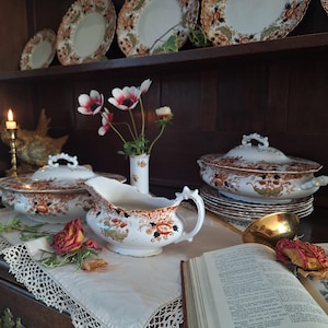 Pair of antique Crown Derby Imari tureens arranged on a lace-covered oak sideboard beneath matching dinner plates, styled with garden anemones, trailing ivy, dried roses and vintage decorative books.