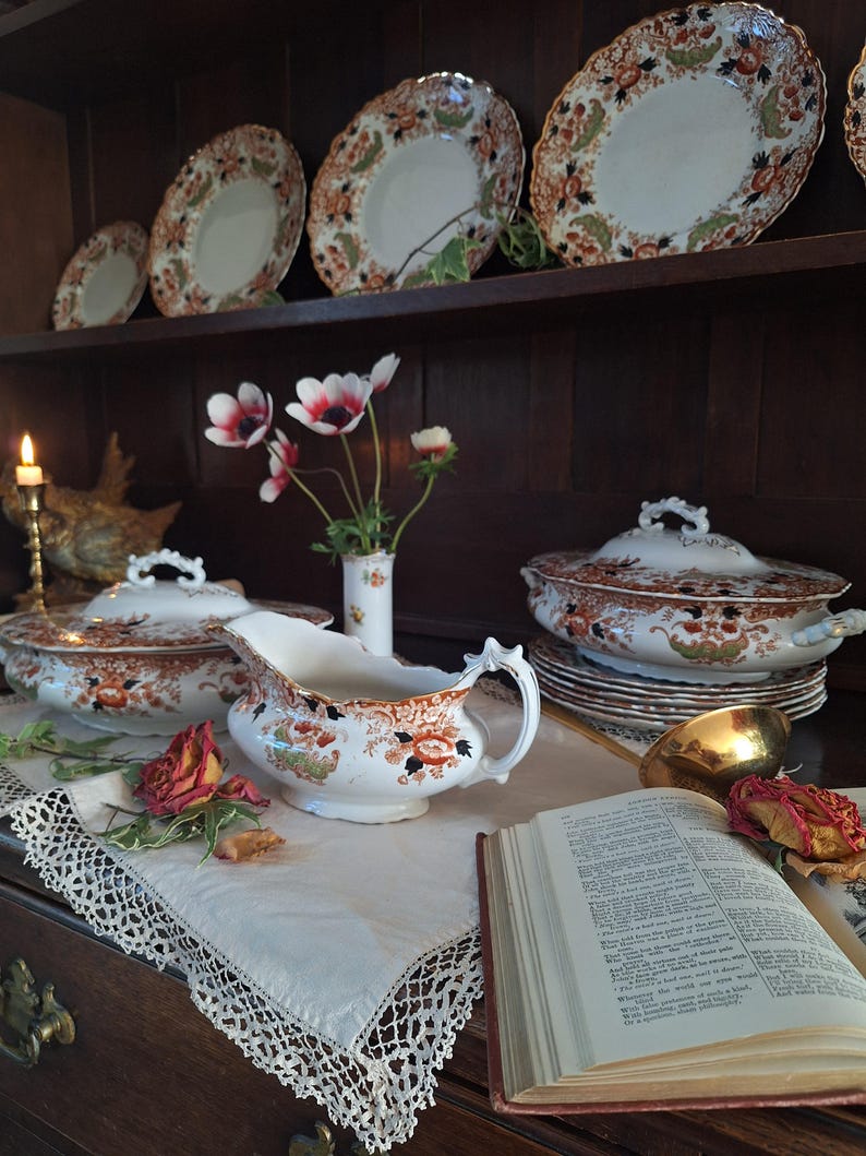Pair of antique Crown Derby Imari tureens arranged on a lace-covered oak sideboard beneath matching dinner plates, styled with garden anemones, trailing ivy, dried roses and vintage decorative books.