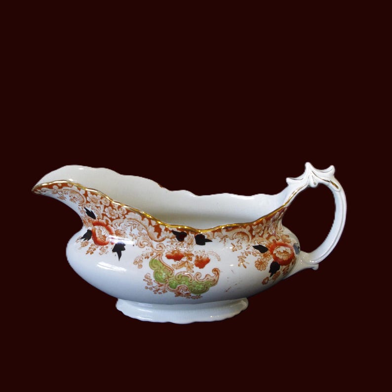 Antique W. Adams Crown Derby Imari Style gravy boat with white base with an orange, green, and black floral pattern. white ceramic gravy boat with a decorative handle and scalloped edge. It has a floral design in orange, green, and black, with gold detailing. A traditional serving piece for sauces and gravies.