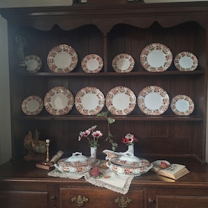 Antique Crown Derby Imari dinnerware displayed on a dark oak dresser with matching tureens, serving pieces, garden flowers, lace linens, brass candlestick and vintage books in an old English country house style interior.