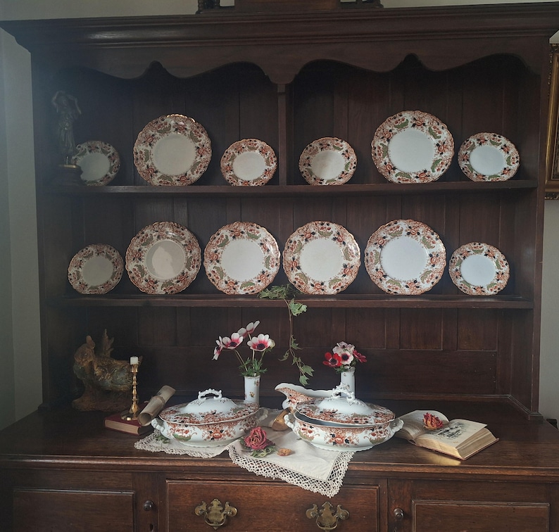 Antique Crown Derby Imari dinnerware displayed on a dark oak dresser with matching tureens, serving pieces, garden flowers, lace linens, brass candlestick and vintage books in an old English country house style interior.