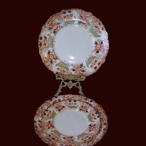 K&ouml;nnte beinhalten: Zwei antike Porzellanteller mit einem Blumendesign in Orange, Gr&uuml;n und Schwarz, pr&auml;sentiert auf einem dekorativen, goldfarbenen St&auml;nder. Die Teller haben einen gewellten Rand und einen goldenen Rand. Die Teller stehen vor einem dunkelroten Hintergrund.