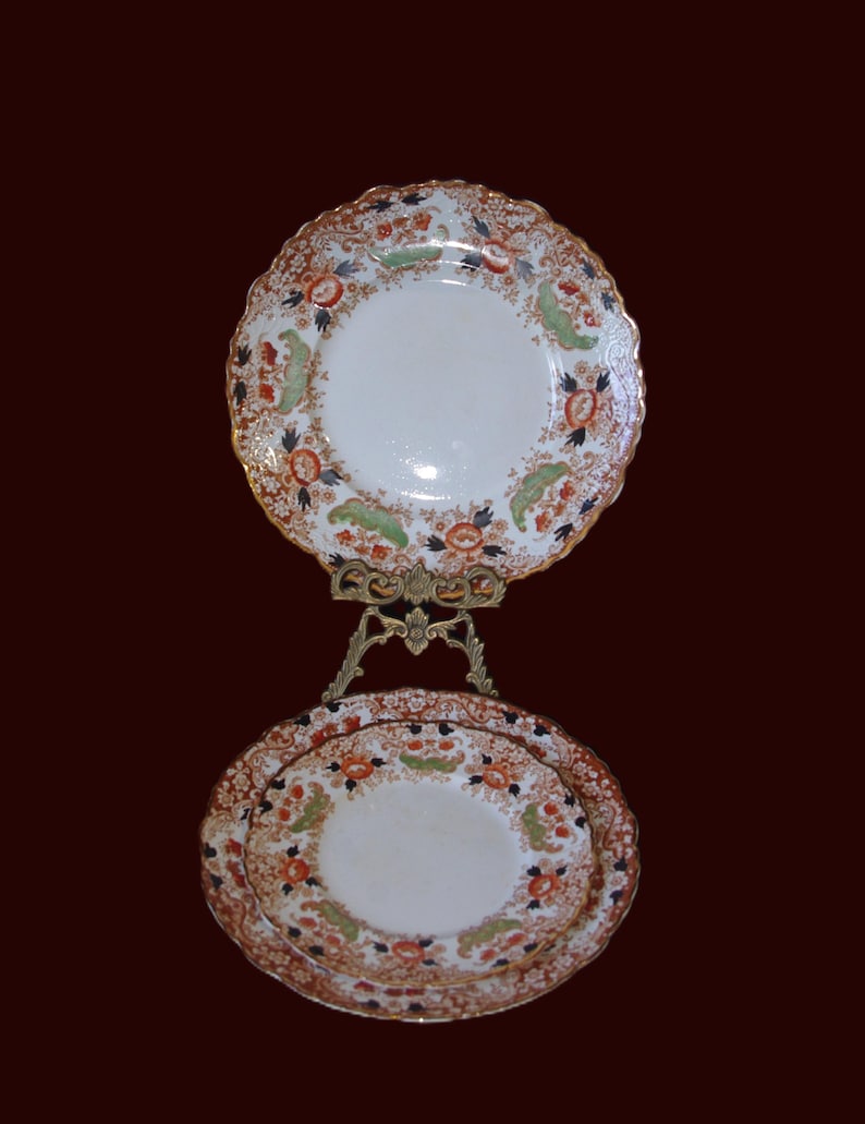 K&ouml;nnte beinhalten: Zwei antike Porzellanteller mit einem Blumendesign in Orange, Gr&uuml;n und Schwarz, pr&auml;sentiert auf einem dekorativen, goldfarbenen St&auml;nder. Die Teller haben einen gewellten Rand und einen goldenen Rand. Die Teller stehen vor einem dunkelroten Hintergrund.