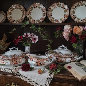 Antique Crown Derby Imari dinnerware displayed on a dark oak dresser with matching tureens, serving pieces, garden flowers, lace linens, brass candlestick and vintage books in an old English country house style interior.
