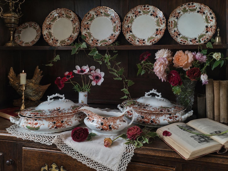 Antique Crown Derby Imari dinnerware displayed on a dark oak dresser with matching tureens, serving pieces, garden flowers, lace linens, brass candlestick and vintage books in an old English country house style interior.