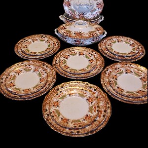 K&ouml;nnte beinhalten: Antikes Geschirr mit einem floralen Muster in Orange, Gr&uuml;n und Braun. Das Set umfasst mehrere Teller, eine Serviersch&uuml;ssel mit Deckel und eine kleinere Schale. Die Teller haben einen goldenen Rand.