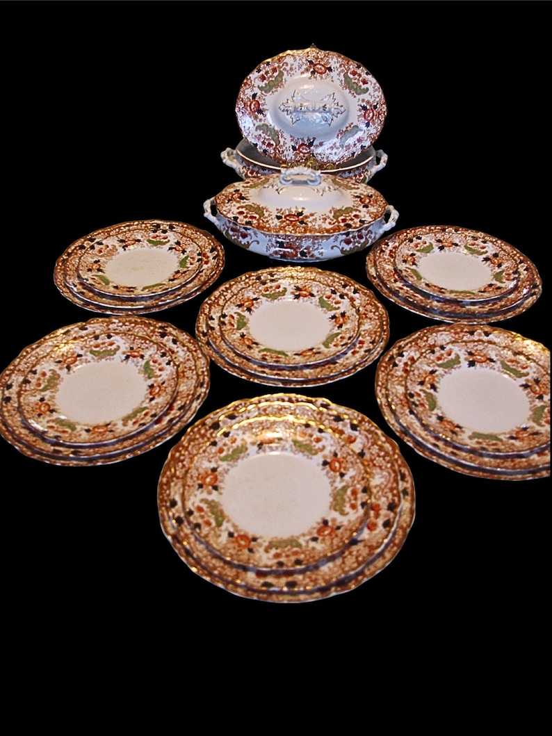K&ouml;nnte beinhalten: Antikes Geschirr mit einem floralen Muster in Orange, Gr&uuml;n und Braun. Das Set umfasst mehrere Teller, eine Serviersch&uuml;ssel mit Deckel und eine kleinere Schale. Die Teller haben einen goldenen Rand.