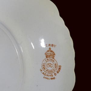 K&ouml;nnte beinhalten: Ein wei&szlig;er Porzellanteller mit gewelltem Rand. Der Teller hat ein braunes Emblem mit den Worten "DERBY", "CROWN SEMI-PORCELAIN", "W. ADAMS & SONS" und "ENGLAND".