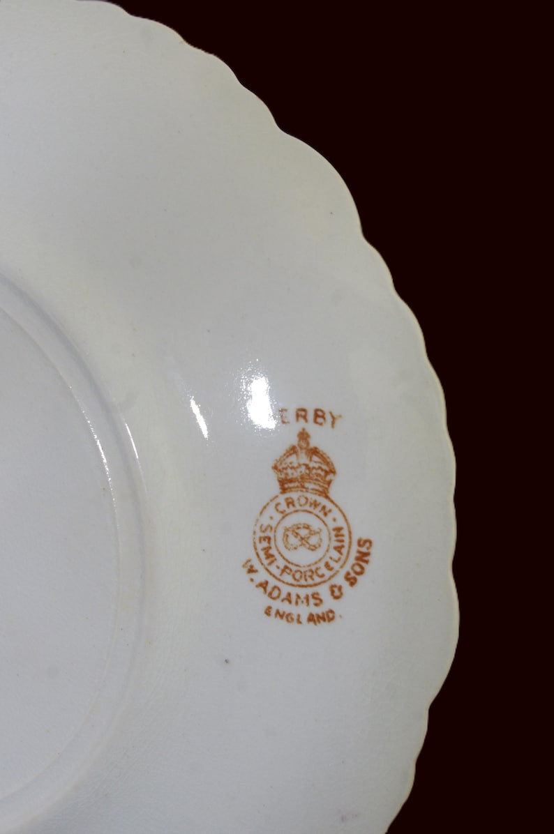 K&ouml;nnte beinhalten: Ein wei&szlig;er Porzellanteller mit gewelltem Rand. Der Teller hat ein braunes Emblem mit den Worten "DERBY", "CROWN SEMI-PORCELAIN", "W. ADAMS & SONS" und "ENGLAND".