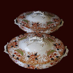 K&ouml;nnte beinhalten: Zwei antike, ovale Servierschalen mit Deckel. Die wei&szlig;en Porzellanschalen weisen ein Blumenmuster in Orange, Gr&uuml;n und Schwarz mit Goldverzierungen auf. Jeder Deckel hat einen dekorativen Griff und Seitengriffe. Der Hintergrund ist dunkelrot.