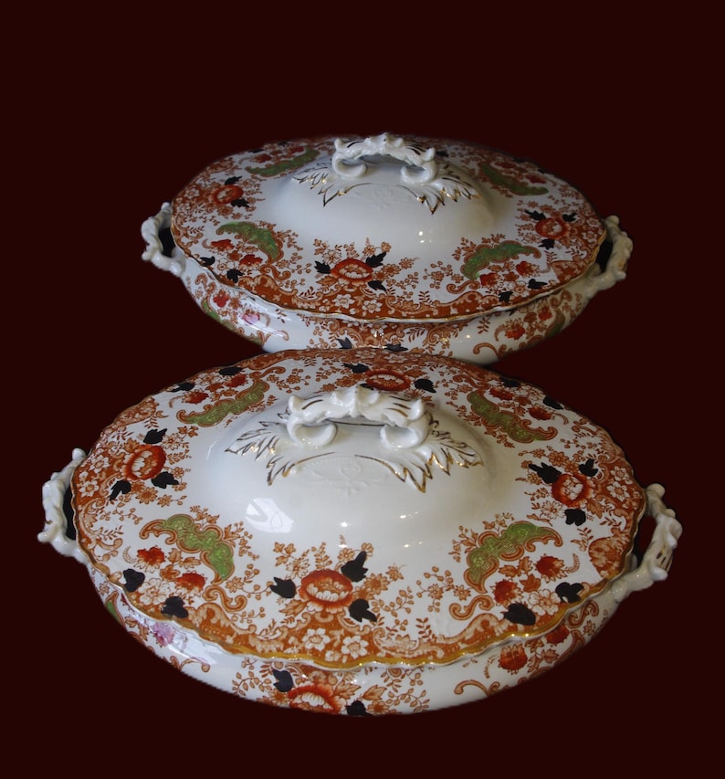 K&ouml;nnte beinhalten: Zwei antike, ovale Servierschalen mit Deckel. Die wei&szlig;en Porzellanschalen weisen ein Blumenmuster in Orange, Gr&uuml;n und Schwarz mit Goldverzierungen auf. Jeder Deckel hat einen dekorativen Griff und Seitengriffe. Der Hintergrund ist dunkelrot.