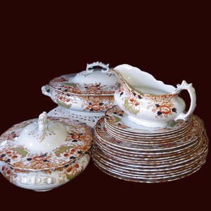 Antique W. Adams Crown Derby Imari Style Dinner Service including a lidded serving dish, a gravy boat, and a stack of plates. The set has a white base with an orange, green, and black floral pattern. The plates have gold rims.