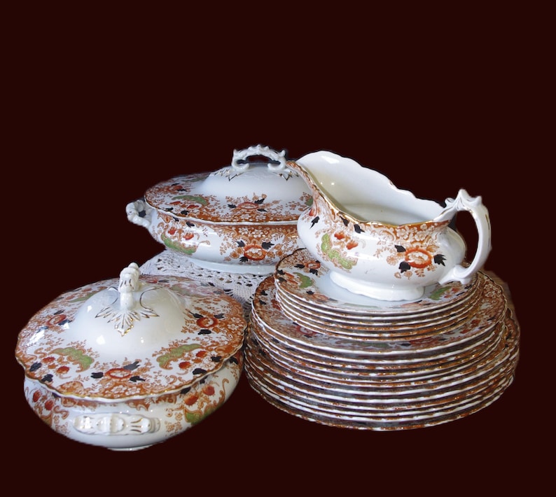 Antique W. Adams Crown Derby Imari Style Dinner Service including a lidded serving dish, a gravy boat, and a stack of plates. The set has a white base with an orange, green, and black floral pattern. The plates have gold rims.