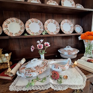 K&ouml;nnte beinhalten: Eine Sammlung antiker Porzellanwaren mit einem floralen Muster in Orange, Braun und Gr&uuml;n. Das Set umfasst Teller, Servierschalen und eine Sauciere, die auf einer Holzkommode und einem Tisch ausgestellt sind. Die Teller haben einen gewellten Rand.