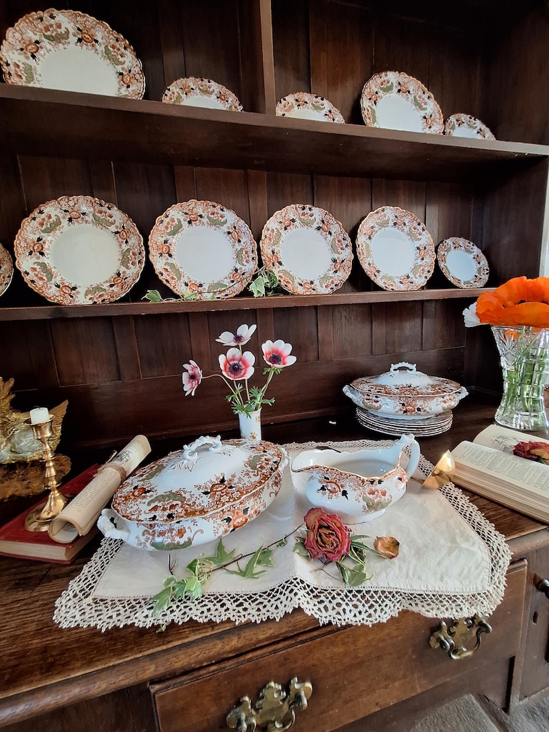 K&ouml;nnte beinhalten: Eine Sammlung antiker Porzellanwaren mit einem floralen Muster in Orange, Braun und Gr&uuml;n. Das Set umfasst Teller, Servierschalen und eine Sauciere, die auf einer Holzkommode und einem Tisch ausgestellt sind. Die Teller haben einen gewellten Rand.