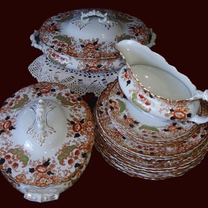 K&ouml;nnte beinhalten: Ein antikes Porzellanservice mit einem floralen Muster in Orange, Gr&uuml;n und Schwarz auf wei&szlig;em Grund. Das Set umfasst Serviersch&uuml;sseln mit Deckel, Teller und eine Sauciere, alle mit Goldverzierungen.