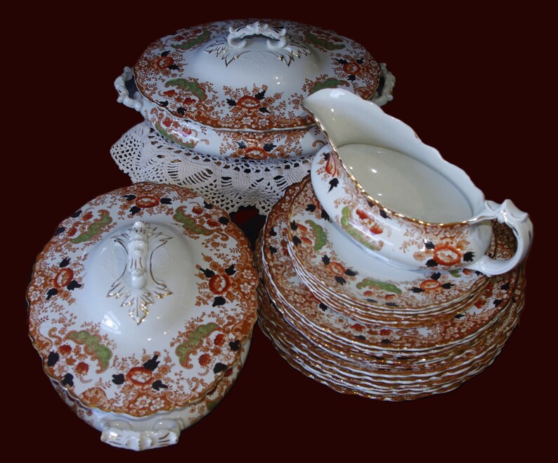 K&ouml;nnte beinhalten: Ein antikes Porzellanservice mit einem floralen Muster in Orange, Gr&uuml;n und Schwarz auf wei&szlig;em Grund. Das Set umfasst Serviersch&uuml;sseln mit Deckel, Teller und eine Sauciere, alle mit Goldverzierungen.