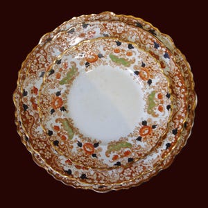 Three Close up from top of Antique W. Adams Crown Derby Imari Style  ceramic plates stacked, featuring a detailed floral design in orange, green, black, and white. The plates have a scalloped edge and gold trim, set against a dark brown background.