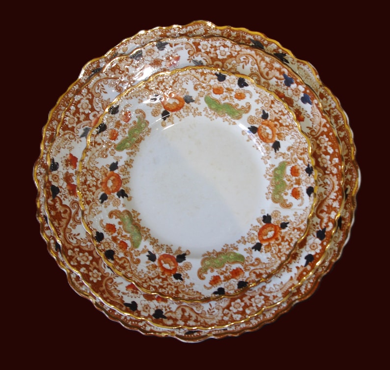 Three Close up from top of Antique W. Adams Crown Derby Imari Style  ceramic plates stacked, featuring a detailed floral design in orange, green, black, and white. The plates have a scalloped edge and gold trim, set against a dark brown background.