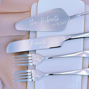 May include: A set of silver cake serving utensils, including a server, knife, and two forks, presented on a white plate with gold edging. The server and knife are engraved with "The Roberts" and the date "10.24.2027". Feathers and a perfume bottle are in the background.