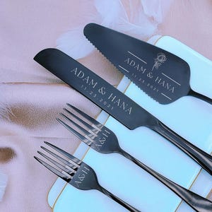 May include: A black wedding cake knife and server set, engraved with "ADAM & HANA" and the date "11.23.2021". Two forks are also included, with "A&H" engraved on the tines. The set is displayed on a white rectangular plate with a gold trim.
