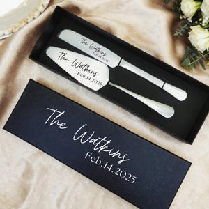 May include: A wedding cake knife and server set in a black box. The set includes a knife and server with the text "The Watkins Feb.14.2025". The box also has the same text. The set is on a beige surface.