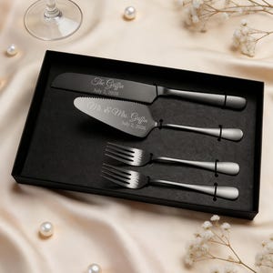 May include: A matte black cake serving set in a black presentation box, including a knife and server engraved with "The Griffin July 2, 2026" and "Mr. & Mrs. Griffin July 2, 2026," plus two matching forks engraved with "Mr." and "Mrs."