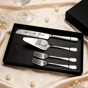 May include: A silver-toned cake serving set in a black box, including a knife and server engraved with Mr. & Mrs. Benak August 29, 2026, and two forks engraved with the initials S and J. The utensils feature smooth, polished handles and metallic finish.