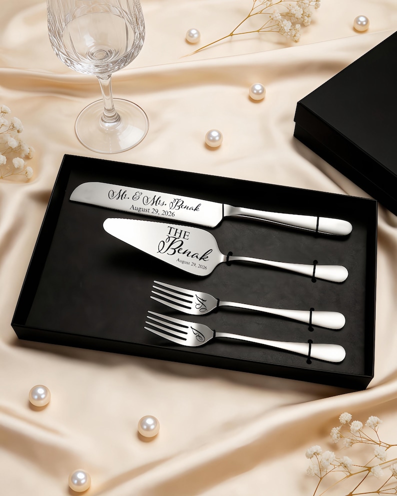 May include: A silver-toned cake serving set in a black box, including a knife and server engraved with Mr. & Mrs. Benak August 29, 2026, and two forks engraved with the initials S and J. The utensils feature smooth, polished handles and metallic finish.