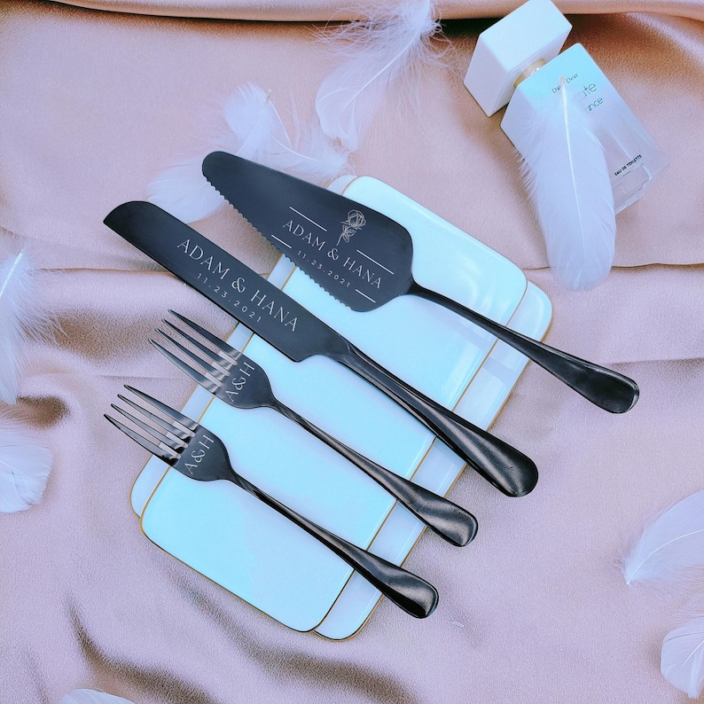 May include: A set of matte black stainless steel cutlery including a cake knife, a cake server, and two forks. The cake server is engraved with ADAM & HANA 11.23.2021 and a rose. The knife reads ADAM & HANA 11.23.2021, and the forks are marked A&H.