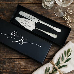 May include: A silver cake knife and server set with a serrated edge, engraved with the text LARA & SASCHA 11.09.2026 and a heart design. The utensils are presented in a black rectangular box featuring the same heart logo on the lid.