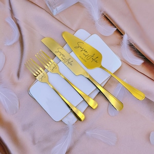 May include: A set of gold-toned stainless steel cutlery including two forks, a knife, and a cake server. The knife and server are engraved with the text Susan and Julia, and the forks are engraved with S&J. The server also features the date 05.08.2025.