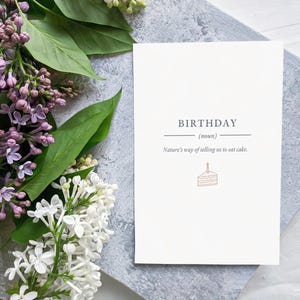 May include: A white birthday card with the word "BIRTHDAY" and the definition "(noun)". The card also has the text "Nature's way of telling us to eat cake." and a cake illustration. Lilac flowers and green leaves are in the background.
