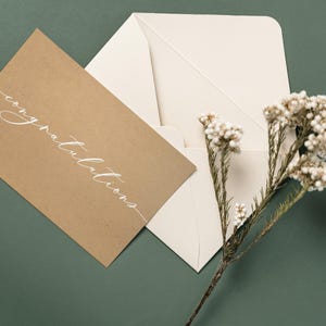 Może przedstawiać: Karta okolicznościowa z papieru typu kraft ze słowem "gratulacje" w eleganckim białym skrypcie, obok otwartej kremowej koperty. Gałązka suszonych białych kwiatów spoczywa na ciemnozielonym tle.