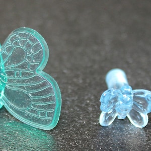 May include: Two plastic butterfly ornaments, one teal blue and one light blue. The teal blue butterfly is larger and has more intricate details.