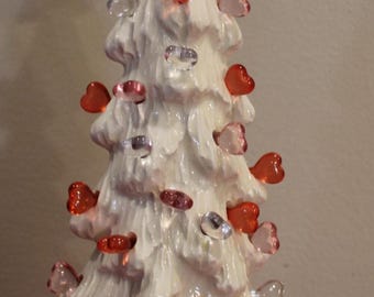 Puede incluir: Un árbol de Navidad de cerámica blanca con acabado brillante, adornado con adornos rojos y transparentes en forma de corazón. Un ángel rojo corona el árbol. Una decoración festiva.