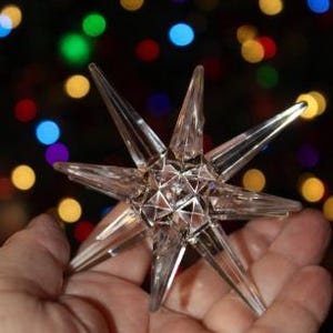 May include: A clear, faceted star-shaped ornament with multiple points, held in a hand. The ornament has a geometric design and is transparent. The background features blurred, colorful lights, suggesting a festive or holiday theme.