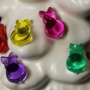 May include: A white ceramic Christmas tree with five colorful bunny-shaped ornaments. The ornaments are purple, green, yellow, and pink.