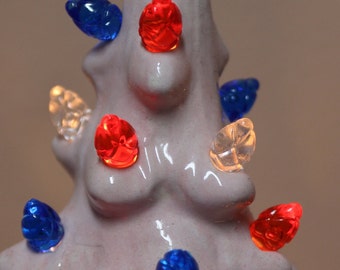 Puede incluir: Un árbol de Navidad de cerámica blanca decorado con bombillas de luz rojas, azules y transparentes. El árbol está adornado con luces de varios colores.