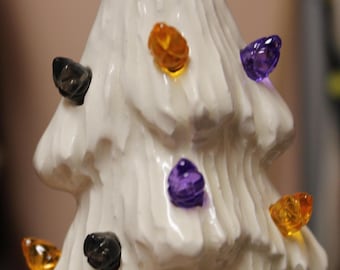 Puede incluir: Un árbol de mesa de cerámica blanca con una superficie texturizada similar a la nieve. El árbol está decorado con pequeñas bombillas de plástico translúcido y multicolor en tonos naranja, morado y negro, insertadas en las ramas.