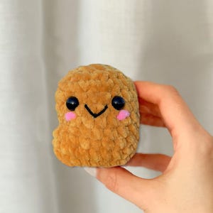 May include: A brown crocheted plush toy shaped like a nugget with a smiling face. The toy has black eyes, a small black mouth, and pink cheeks.
