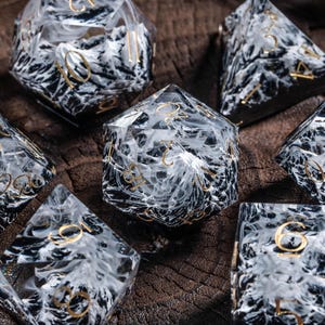 May include: A set of seven polyhedral dice, featuring a black and white marbled pattern with gold numerals. The dice are presented in a wooden bowl. Suitable for role-playing games and other tabletop activities.