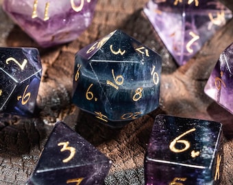 May include: A collection of seven multi-sided dice in shades of purple and blue, with gold numbering. The dice are arranged in a wooden bowl, showcasing their geometric shapes and intricate details. Ideal for tabletop games.