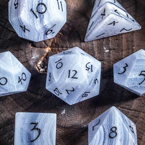 Może przedstawiać: Zestaw siedmiu niebiesko-białych kości do gier RPG. Kości mają różne kształty, w tym kostkę, piramidę i wielościany, każdy z czarnymi liczbami. Kości są ułożone na ciemnej drewnianej powierzchni.
