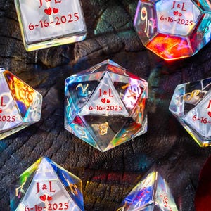 May include: A collection of iridescent, geometric glass objects, each inscribed with "J&L", two hearts, and "6-16-2025". The objects are various polyhedral shapes, including a cube, a dodecahedron, and a pyramid. The objects are displayed on a dark, textured surface.