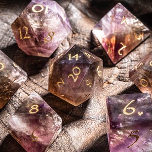 Pode incluir: Conjunto de sete dados poliédricos em tons de roxo, âmbar e transparente, com numeração dourada. Os dados são exibidos em uma tigela de madeira rústica. Os dados são para jogos de RPG.