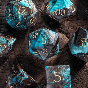 May include: Set of seven blue and black gemstone dice with gold numbers. The dice are on a dark brown wooden surface.