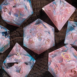 Puede incluir: Juego de siete dados poliédricos para juegos de mesa. Los dados son transparentes con un diseño arremolinado rosa y azul y numeración dorada. Los dados se muestran sobre una superficie de madera oscura.